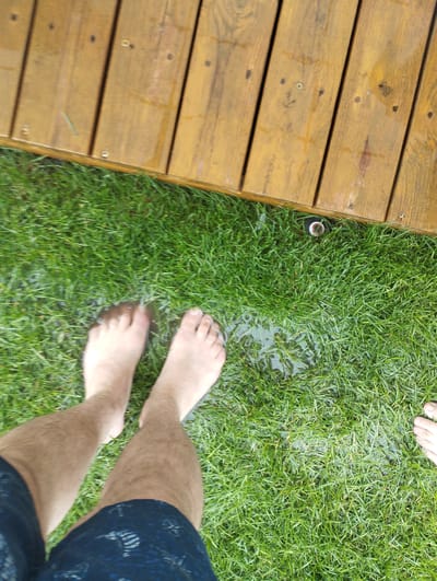This is a top-down, close-up shot of a person's bare feet standing on wet grass, next to a wooden deck. The grass appears to be recently watered, with several small puddles visible. The wooden deck is made of planks and appears to be an outdoor structure, possibly a patio or part of a house. A sprinkler head is partially visible on the grass near the edge of the deck. The image is taken from a high angle, looking down at the feet and grass. The lighting suggests it is daytime, and the wet grass implies it might be raining or the sprinklers have been on recently. There are no visible people other than the legs and feet of the person taking the photo, and no visible text. The scene is set in an outdoor environment, possibly a backyard or garden in Romanivka, Ukraine. The overall mood is casual and relaxed, suggesting outdoor recreation or simply enjoying a moment outside.