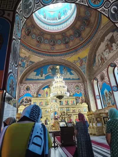 The photograph depicts the interior of a richly decorated Orthodox church, likely located in Ocnița, Moldova. The scene is dominated by intricate frescoes and religious iconography adorning the walls and dome. A large, ornate chandelier hangs from the ceiling, illuminating the gilded iconostasis, which is a screen separating the sanctuary from the nave, filled with religious paintings. Several individuals, primarily women, are visible from behind, facing the iconostasis, suggesting they are participating in a religious service or visiting the church. They are dressed in conservative attire, with headscarves common among them. The architectural style is characteristic of Eastern Orthodox churches, with a central dome and arched doorways. The overall atmosphere is one of solemnity and reverence, typical of a place of worship.