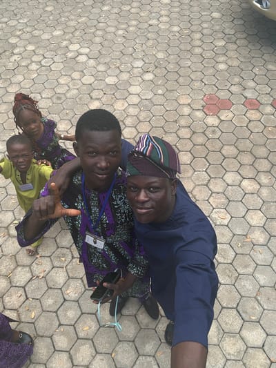In Itoga, Nigeria, two young men, likely teenagers or young adults, are the primary subjects in the foreground, positioned for a selfie. The man on the left, wearing a purple and black patterned traditional outfit with a lanyard around his neck, is holding a smartphone and appears to be making a "L" sign with his left hand. The man on the right, wearing a blue traditional outfit and a multi-colored striped cap, is also looking towards the camera. Two younger children, a girl with braided hair wearing a purple outfit and a boy in a light green outfit, are visible behind the men, looking at the camera with somewhat uncertain expressions. They are all standing on a paved outdoor area composed of hexagonal concrete blocks, with a few fallen leaves scattered on the ground. The overall lighting suggests it is daytime.