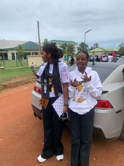 Two young women stand beside a silver car on a dirt road. The woman on the left, wearing a white t-shirt with graphics and a black graduation sash, holds a black marker and gestures with her left hand. The woman on the right, in a white hoodie with a yellow bird graphic, makes a hand gesture with both hands. In the background, a large, modern building complex with a green roof is visible, possibly a school or administrative center, with a crowd of people gathered in front of it. Power lines and a wooden utility pole are also present. The sky is overcast. The location is identified as South Ropp, Nigeria.