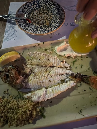 This is a top-down, close-up photograph of a partially consumed meal on a pale green rectangular plate. The plate is set on a wooden surface, possibly a table, with a blue and white patterned dish and a patterned placemat visible in the upper portion of the frame.
The main subject is a cooked fish, mostly deboned, with visible white flesh and a charred head and tail. The fish is seasoned with herbs and appears to be drizzled with olive oil. To the left of the fish, a wedge of lemon rests on the plate. In the lower left corner, there is a pile of what appears to be cooked grains, possibly bulgur or couscous.
To the right of the fish, on the plate, is a dollop of white sauce or dip, possibly tahini or a yogurt-based sauce. A hand is visible in the upper right corner, holding a small glass bottle with a spout, from which a yellow liquid, likely olive oil or a dressing, is being poured over the meal. The patterned dish in the background is partially eaten, with remnants of food and a fork and knife resting on it. The placemat beneath the dishes has a faded drawing of a plant and some text, partially obscured.
The lighting suggests an indoor setting, possibly a restaurant in Asprovalta, Greece, given the context. The overall impression is of a finished or nearly finished meal, with elements of preparation and consumption visible.