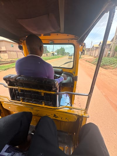 The image shows the interior of a yellow auto-rickshaw, commonly known as a "Keke Napep," in motion. The camera is positioned inside the vehicle, looking forward and to the side. The main subject is the driver, a man wearing a purple shirt and a blue arm sleeve, with his hands on the steering wheel. He is looking forward, presumably at the road ahead.
The auto-rickshaw appears to be on a dirt road, with dry, reddish-brown soil. On either side of the road, there are signs of settlement with buildings and vegetation. In the background, lush green foliage can be seen, suggesting a tropical or sub-tropical environment. The sky is cloudy, indicating it might be a warm day, but not necessarily sunny. The overall scene depicts everyday transportation in an urban or semi-urban area of Enugu, Nigeria.
Visible objects include the driver's seat, the steering wheel, and parts of the auto-rickshaw's yellow exterior. There is a side mirror visible. On the seat behind the driver, there is a blue and white object with some text on it, partially obscured. The interior of the auto-rickshaw shows some wear and tear, with parts of the black seat upholstery visibly damaged.
The image captures a moment of travel, with the motion of the vehicle implied by the slight blur of the background. The focus is on the driver and the immediate surroundings of the auto-rickshaw, providing a glimpse into local transportation and daily life.