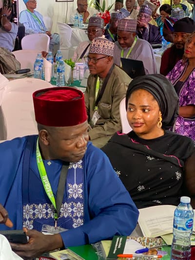 The image shows a conference setting in what appears to be Kwarau, Nigeria. Several people are seated at tables, some wearing traditional attire. A woman in a black headscarf and dress with floral accents is seated at the foreground of the table. A man in a blue outfit and a red hat is sleeping, with an accreditation lanyard around his neck labeled "ACReSAL". A book titled "Kaduna ACReSAL Implementation in Pictures" is visible on the table next to a bottle of water.