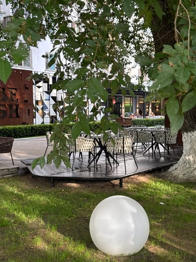 This image depicts an outdoor seating area of a restaurant or cafe, likely in Pavlodar, Kazakhstan. The scene is viewed through the leaves of a tree in the foreground, which are slightly out of focus and cast dappled light on the surroundings.
The main subject is a raised wooden deck furnished with numerous chairs and tables. The chairs appear to be a modern design with woven seats and dark metal frames. The tables are also dark and have a sleek, contemporary look. Behind the seating area, a building with a distinctive facade featuring geometric patterns and glass windows is visible. Some people are seated at tables inside the building, enjoying a meal or drinks.
To the right of the deck, a large, spherical white light fixture sits on a patch of well-maintained green grass. The lighting suggests it is daytime, possibly late afternoon or early evening, as there are shadows cast on the grass. The overall atmosphere appears relaxed and inviting, suggesting a pleasant outdoor dining experience. The presence of greenery and the open-air setting indicate a temperate climate. No visible text is present in the image.