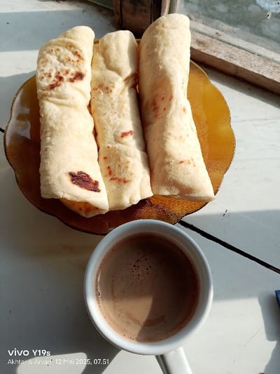 Three flatbreads are arranged on a brown, scalloped plate. The plate is on a white surface next to a white cup of coffee. The flatbreads are slightly browned from cooking. The coffee is dark brown and has a frothy top. The scene is in a home in Anjatan, Indonesia, at the coordinates -6.419 latitude, 107.978 longitude. There is a window in the background with a wood frame.  The image was taken on May 12, 2023 at 07:51.  The image was taken with a Vivo Y19s phone.  There are no people in the image.