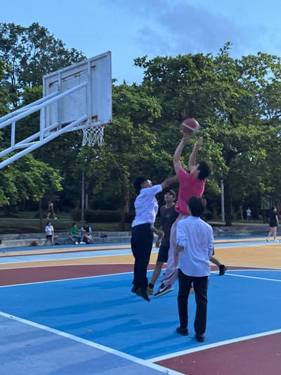 The image captures a dynamic moment on a brightly colored outdoor basketball court. The main subjects are a group of young people engaged in a playful basketball activity. The central figure, wearing a pink t-shirt, is mid-air, reaching out to catch a basketball. Another person in a white shirt is positioned to their left, also appearing to be in motion. Below them, two more individuals are visible, one in a white top and black pants, and another further down in dark clothing, suggesting a coordinated or spontaneous game.

The setting is an outdoor court with distinct blue, red, and yellow lanes, characteristic of a recreational sports facility. In the background, lush green trees provide a natural backdrop, hinting at a sunny day with clear skies. A basketball hoop and backboard are clearly visible on the left side of the frame. The overall atmosphere appears energetic and joyful, with the subjects appearing focused and engaged in their activity. The image appears to have been taken during daylight, likely late afternoon given the light and shadows.

There is no visible text in the image. The context provided places the scene in Phuket City Municipality, Thailand.