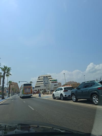 The image depicts a sunny day on a street in San Javier, Spain. The view is from inside a vehicle, looking forward at a multi-lane road. In the foreground, the hood of the vehicle is visible, reflecting the sky and surrounding environment.

On the right side of the road, two cars are parked: a dark green SUV and a white SUV. Further down the road, on the left, a white bus with colorful advertisements is driving away from the camera. The bus has "TOROS DE MURCIA" and "SEPTIEMBRE 2023" printed on its rear.

In the background, a distinctive, stepped pyramid-shaped building is prominently visible. To the left of the pyramid building, other buildings and palm trees line the street. Traffic lights are visible, with one showing a green light. Pedestrians can be seen walking on the sidewalk near the pyramid building. The sky is mostly clear blue with some white clouds. Signage is present, indicating directions to "GRAN VIA" and "playa del Galán". The environment suggests a coastal or resort town.