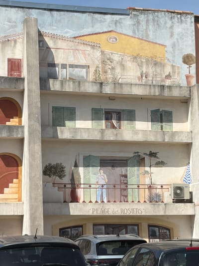 This is a photograph of a mural painted on the side of a building in Mercurol-Veaunes, France. The mural depicts a multi-story building with balconies and windows. On the top level, a yellow building with a red-tiled roof is visible, with potted plants and trees on a balcony. The middle levels show windows with green shutters and balconies. The lower levels depict a balcony with a man standing and looking out. Below the mural, there are store entrances and parked cars. The mural is colorful and detailed, giving the impression of a Mediterranean street scene.