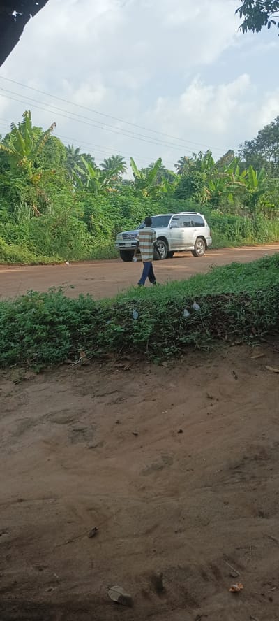 A daytime scene shows a rural dirt road in Amauzu, Nigeria. Lush green vegetation lines both sides of the road, including banana trees and other tropical foliage. An older model silver Lexus SUV is parked on the side of the road. A man in a striped shirt and dark pants is walking on the road, away from the vehicle. Scattered trash is visible along the side of the road in the foreground. Overhead power lines traverse the scene. The sky is cloudy with patches of blue.