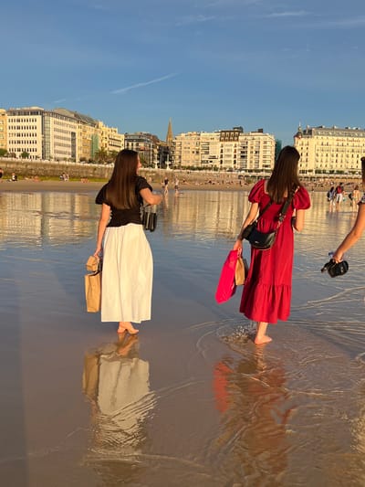 Two women walk along the shore of La Concha Beach in San Sebastián, Spain, near the promenade. The woman on the left wears a black top and a long white skirt, carrying a brown paper shopping bag and a smaller beige bag. The woman on the right wears a red dress and a black crossbody bag, holding a bright pink shopping bag and a brown paper shopping bag. Both women are barefoot, with shallow water lapping around their feet. In the background, the elegant buildings lining the promenade are visible, reflecting in the wet sand under a clear blue sky. Several other people are scattered on the beach and in the water, with more buildings and a church spire further in the distance. The scene is illuminated by the warm light of either a sunrise or sunset.