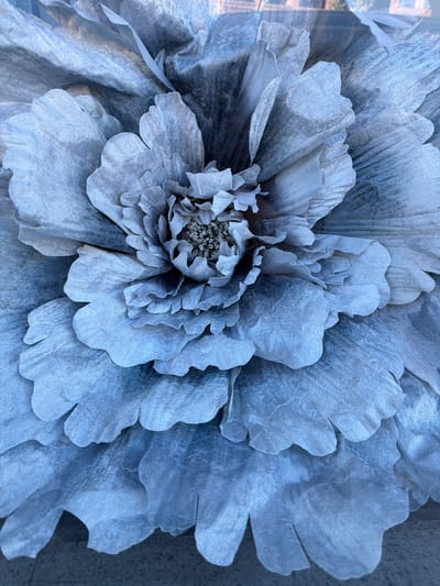 A close-up shot of a large, artificial flower, likely a decorative piece. The flower is predominantly silver or light gray, with intricate petal arrangements leading to a densely packed center. The texture of the flower appears to be fabric-like. The background is blurry and indeterminate. There is no indication of the location in Azuqueca De Henares, Spain, or any information about people, actions, or interactions in the provided image.