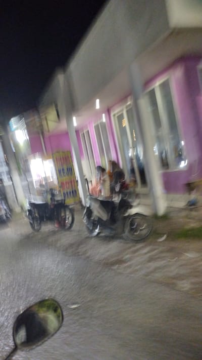 This media file depicts a street scene at night in Bangsri, Indonesia. The image is blurred due to motion, likely taken from a moving vehicle.
The subjects include several motorcycles. On the left, a motorcycle with red accents on its wheels is parked near a stall or shop with a yellow sign. The sign has text on it, appearing to read "PISANG KEJU ROTI BAKAR". To the right, another motorcycle is visible, with two people seated on it. One person is wearing an orange shirt, and the other is wearing a brown hijab. A person in a light-colored shirt is also near the motorcycles. The background features a building with a purple facade and large windows, illuminated by interior lights. The ground appears to be wet or reflective, suggesting recent rain or a damp surface.
The overall scene suggests a bustling street environment, possibly a night market or roadside food stalls. The time of day is clearly night, indicated by the darkness in the upper portion of the image and the artificial lighting. There are no strong indicators of weather, other than the possibility of dampness on the ground. The motion blur conveys a sense of movement and transience.
