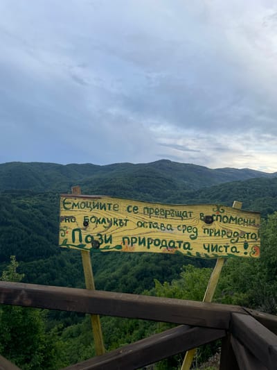 This media file shows a close-up of a wooden sign with text written in Cyrillic, set against a backdrop of a lush, green mountainous landscape under a cloudy sky. The sign is weathered and painted yellow with green and red lettering, featuring some decorative floral elements. The text on the sign reads, "Емоциите се превръщат воспомени. но Боклукът остава сред природата! • Пази природата чиста." This translates to "Emotions turn into memories. but the trash remains in nature! • Keep nature clean." The sign is attached to a wooden post, which is part of a larger wooden structure, possibly a fence or viewing platform, visible on the right side of the frame. The mountains are covered in dense forest, suggesting a natural, perhaps rural or wilderness, setting. The sky is overcast with some patches of brighter clouds, indicating it could be daytime, possibly morning or afternoon, with mixed weather conditions. The overall scene evokes a sense of environmental awareness and responsibility, highlighting the beauty of nature and the importance of preserving it. There is also a faint inscription "FAP" on one of the wooden beams. The location is identified as Delchevo, Bulgaria.