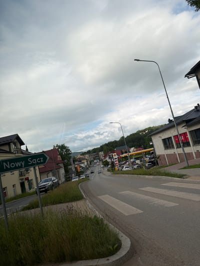 The image shows a view of a street in Limanowa, Poland. A green directional sign with white text reads "Nowy Sącz". The street is paved and has a pedestrian crossing marked with white stripes. Several cars are visible on the road, and some are parked. Buildings line the street, including what appears to be a gas station with a Shell logo and pumps. There are streetlights and overhead wires. The sky is cloudy. Lush green grass and vegetation grow along the roadside and on a median. The overall scene depicts a typical urban or suburban street environment.