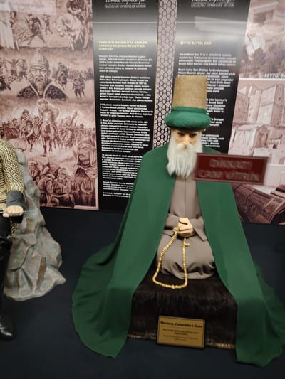 This image captures a museum exhibit featuring wax figures and informational displays in Eskisehir, Turkey. The central subject is a lifelike mannequin depicting Mevlana Celaleddin-i Rumi, identified by a plaque at its base. He is seated cross-legged, adorned in a dark green turban and a flowing dark green cloak over a beige robe, holding a string of golden prayer beads (tasbih). His face features a prominent long white beard.
To the left, part of another mannequin is visible, dressed in a grey metallic chainmail-like tunic and dark trousers, with a hand resting on a textured base.
The background consists of large, sepia-toned informational panels. One panel details "TÜRKLER'İN ANADOLU'YA AKINLARI ANADOLU SELÇUKLU DEVLETİ'NİN KURULUŞU" (Invasions of Turks to Anatolia, Establishment of the Anatolian Seljuk State), providing historical context about events like the Malazgirt Victory, the First Crusade, and the significance of Eskişehir. It features illustrations of warriors, battles, and historical figures. Another panel focuses on "SEYYID BATTAL GAZI," describing his life and contributions. Both panels bear the title "BALMUMU HEYKELLER MÜZESİ" (Wax Sculptures Museum). A blurred red sign is visible, which appears to warn "DİKKAT CAM KIRILIR" (Attention, glass breaks).
The scene is an indoor museum setting, designed to educate visitors about historical and cultural figures and events relevant to the region. The figures are static displays, presenting historical personalities.