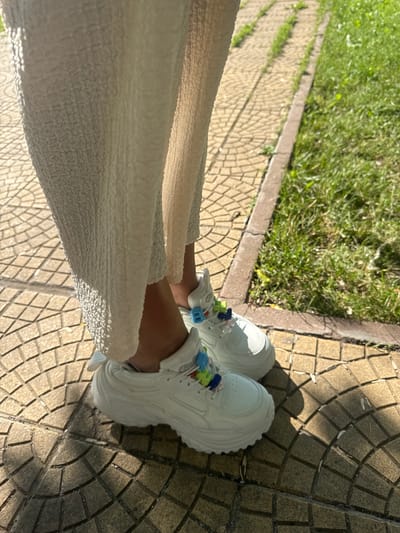 In Kostinbrod, Bulgaria, a person stands on a sun-dappled, patterned concrete sidewalk adjacent to a strip of green grass. The individual is wearing a textured, white, ankle-length garment and white sneakers adorned with colorful lace charms. The shadow of the person falls across the sidewalk.