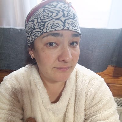 This is a close-up, head-and-shoulders selfie of a woman taken indoors. She is wearing a cream-colored, fuzzy robe and a patterned headscarf with a rose motif and a small, decorative embellishment. Her expression is neutral, with a slight hint of a smile. She has dark brown eyes and dark hair visible around her face and peeking out from under the headscarf. She is wearing a small, sparkly hoop earring in her visible left ear. The background suggests an indoor setting, possibly a home, with hints of curtains, a textured grey surface, and wooden paneling. The lighting is soft, and the overall atmosphere is casual and relaxed. There is no visible text in the image. The location context is Podlubovo, Russia.