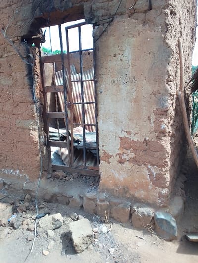 The image shows the exterior of a damaged building in Kwal, Nigeria, located at coordinates 9.803 latitude, 8.646 longitude. The building is constructed of mud brick and has a doorway with a metal gate. The gate is partially open and there are several pieces of corrugated metal lying inside the doorway. There is a wire running along the wall and a wooden stick leaning against the building. The wall is cracked and damaged, and there is some debris on the ground. The letters “WZ” are written on the wall. The image appears to be taken from a street-level perspective.