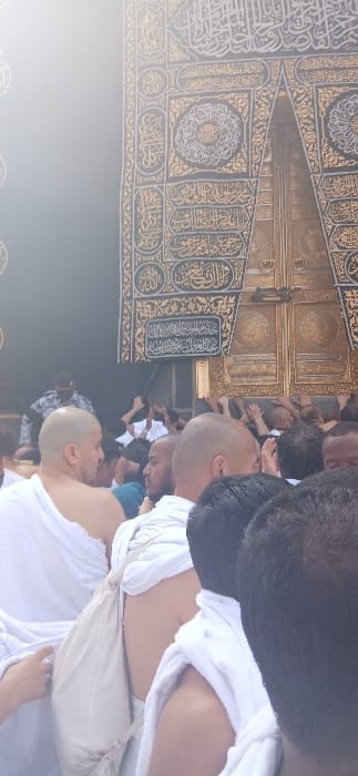 The image displays a dense gathering of pilgrims at the Kaaba in Makkah Al Mukarramah, Saudi Arabia. The upper right section of the frame features a portion of the Kaaba's black Kiswah, elaborately embroidered with golden Arabic calligraphy, next to the ornate golden door, identified as Bab Al-Tawbah or Bab Ibrahim. In the foreground and middle ground, numerous individuals, predominantly men, are visible. Many are dressed in white, seamless Ihram garments, characteristic of Hajj or Umrah rituals. Several men in the crowd are bald, suggesting the completion of the *halq* ritual. The crowd is tightly packed, with multiple hands extended and raised towards the Kaaba and its door, indicating a collective effort to touch or pray near the sacred structure. One pilgrim in the immediate foreground carries a light-colored shoulder bag. The scene conveys a moment of intense religious devotion and significant human congestion during pilgrimage.