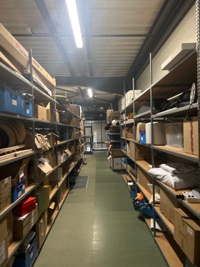 The image depicts an interior view of a storage facility located in Reichstett, France. The scene is a long, narrow aisle, approximately 2-3 meters wide, flanked on both sides by multi-tiered metal industrial shelving units extending into the background. The shelving is uniformly filled with various items, primarily brown cardboard boxes of differing sizes, blue plastic storage bins, and rolled materials. On the upper right shelving unit, a circular, reflective object is visible. On a lower right shelf, several bundles of white or light-grey fabric are stacked. The floor is a solid, light green industrial surface, with a dark rectangular mat positioned on the left side of the aisle in the foreground. Overhead, the ceiling is a corrugated metal structure, from which multiple fluorescent strip lights are suspended, providing bright, uniform illumination. At the far end of the aisle, a doorway with a glass panel leads to another area, and a green exit sign is mounted above it. No individuals are present, and no actions or interactions are depicted within the frame.