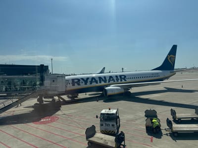 The image depicts a Ryanair airplane parked at an airport gate, likely Barcelona-El Prat Airport in El Prat de Llobregat, Spain, based on the presence of a jet bridge and airport infrastructure. The airplane, with its distinctive blue and yellow livery, is positioned on the tarmac, with its engines visible. A jet bridge connects to the aircraft's fuselage, and a set of stairs leads down from the jet bridge to the ground. Several airport service vehicles are present, including a tug and baggage carts. One individual, wearing a high-visibility vest, is seen near a baggage cart, appearing to be loading or unloading luggage. The scene is set under a clear blue sky with some scattered clouds, suggesting a sunny day. The ground is paved with concrete and marked with yellow and red lines, typical of airport taxiways and apron areas. In the background, other aircraft and airport buildings are faintly visible.