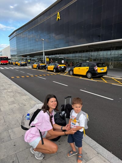 A woman and a young boy are on a sidewalk in front of an airport terminal building in El Prat de Llobregat, Spain. The woman is crouching and looking to her left with a smile, while the boy stands next to her, looking forward. Both are wearing backpacks. The woman has on a pink t-shirt and denim shorts. The boy is wearing a white t-shirt with a graphic print and blue shorts. In the background, several yellow and black taxis are parked in a line along the curb. A bus is visible on the left. The airport terminal has a modern facade with large, dark glass windows and a prominent yellow "A" logo. The sky is partly cloudy.