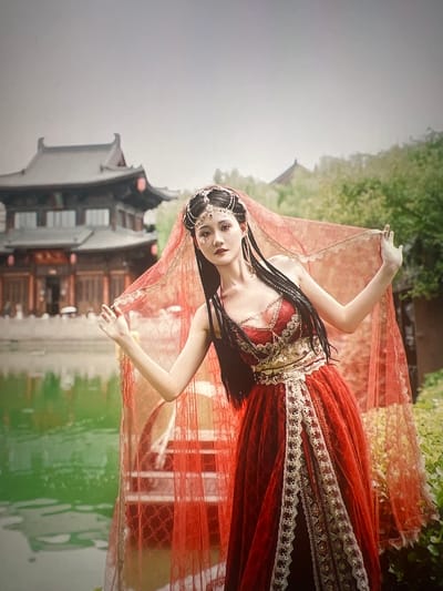 A young woman with dark braided hair adorned with a jeweled headband and facial glitter poses outdoors, dressed in a elaborate red and gold costume. Her dress features intricate gold embroidery and layered red fabric, with a sheer red veil draped over her head and shoulders. She is positioned in front of a traditional Chinese-style building, likely in Liandu District, China. The building has dark wooden elements and distinctive rooflines, and it overlooks a body of green water. The scene appears to be set during the daytime, with soft lighting suggesting overcast weather or shade from surrounding greenery. The woman's expression is serene and contemplative.