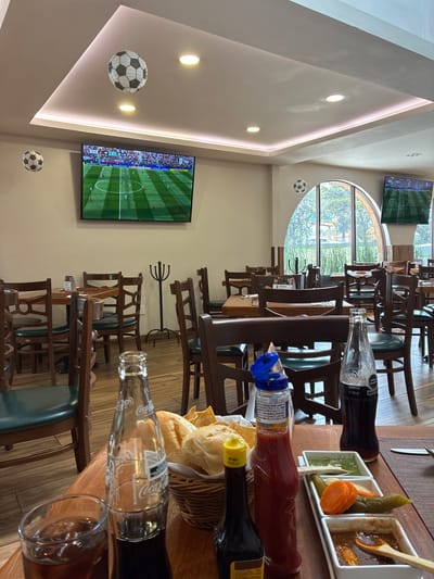 The image shows the interior of a restaurant, likely a sports bar, as evidenced by multiple televisions displaying a soccer match and soccer ball decorations on the ceiling. The restaurant features wooden tables and chairs, a wooden floor, and an arched window with a view of trees and outdoor structures. On one of the tables, a glass of iced beverage, a bottle of Coca-Cola, a basket of bread and chips, a bottle of ketchup, and small dishes of sauces and pickled vegetables are visible. The scene appears to be set for a meal. The location is identified as being in Cuautitlán Izcalli, Mexico.