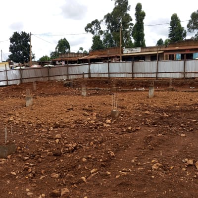 This media file captures a construction site under an overcast sky, likely in a semi-urban or rural area of Borabu Chitago ward, Kenya. The foreground and midground consist of a large, open plot of reddish-brown, uneven earth scattered with numerous rocks and small debris. Several concrete footings with protruding rebar reinforcements are visible across the site, indicating the foundational stage of a new building project.
In the background, a temporary fence constructed from corrugated metal sheets supported by wooden posts delineates the perimeter of the site. Beyond this fence, a row of simple, single-story buildings with corrugated iron roofs can be seen. These structures appear to be local shops or residences. Prominently displayed on one of these buildings is the text "UTUMISHI CYBER & SALON," suggesting the types of businesses operating nearby. Tall, leafy trees are scattered behind and around these buildings, reaching towards the light grey, cloudy sky. No people are visible in the image. The scene depicts a quiet moment in the early development phase of construction.