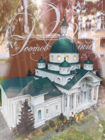 This is a close-up, eye-level, exterior, medium shot of a detailed architectural model of the Church of Elijah the Prophet in Yaroslavl, Russia. The model is made of white plaster with green, sloped roofs and dark green, conical trees. The church features a large central dome with four smaller domes surrounding it, all topped with gold crosses. There are small, round windows on the domes, some of which are illuminated. The facade of the church is adorned with columns and intricate detailing. In front of the church, a procession of miniature figures dressed in yellow and black robes is depicted, suggesting a religious event. The figures are arranged in a line, moving towards the church. The model is situated in what appears to be a display case, as evidenced by reflections on the surface, obscuring some background details. The lighting is soft, with a slight lens flare visible in the upper right quadrant. The overall impression is of a meticulously crafted replica of a significant religious structure.