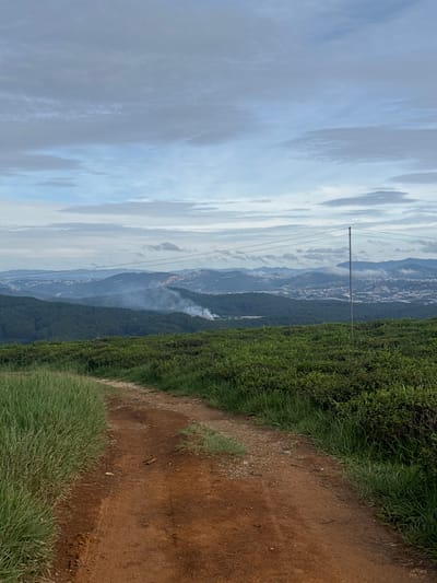 A landscape image shows a dirt path with grass to the side, and green foliage in the background to the left, with a view of a city or town in the distance. The sky is overcast. There is what appears to be smoke in the distance. A metal pole with wires can be seen on the left side of the image. The location is Phát Chi, Vietnam.