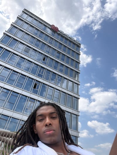 A ground-level medium shot features a person with dreadlocks, partially obscured by the camera angle. Behind them, a tall modern building rises against a cloudy sky. The building's facade is predominantly glass with metallic accents, reflecting the sky. At the top of the building, visible signage with a red logo and what appears to be Chinese characters indicates that it is the Ramada Plaza Hangzhou Linping District. The setting appears to be outdoors during daylight.