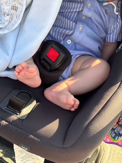 The image shows the lower body of a baby secured in a car seat. The baby is wearing a blue and white striped shirt and appears to be in a stroller or vehicle. The car seat is black with a red buckle. The baby's feet are visible and appear to be resting on the car seat. There is a light blue blanket partially covering the baby. The scene appears to be outdoors, with sunlight casting shadows on the car seat and baby. The image does not contain any information that would allow for the determination of its location in Chorrillos, Peru.