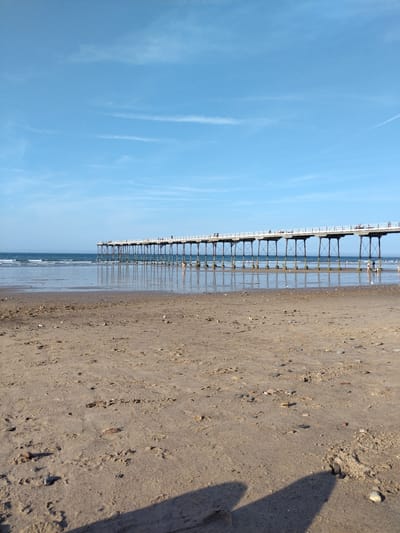 The image shows a view of Saltburn Pier and the beach in Saltburn by the Sea, United Kingdom. The pier, a long, elevated structure with railings, stretches horizontally across the frame, supported by numerous dark, slender pillars. Several indistinct figures are visible walking on the pier. The sea is calm, with gentle waves lapping at the shore. The foreground is a sandy beach with footprints and scattered pebbles. The sky is a clear blue with a few wispy white clouds. The lighting suggests it is daytime, likely in the afternoon, given the shadows cast on the sand.