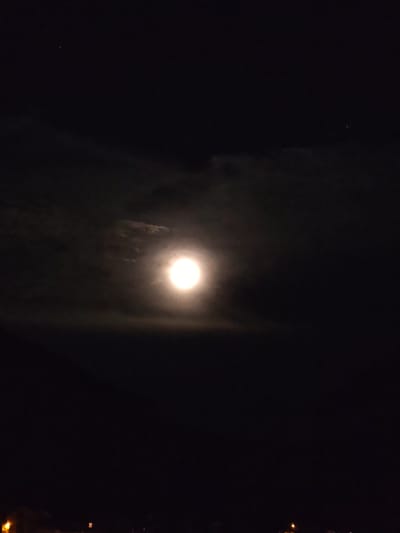 This image captures a nighttime scene in Schwangau, Germany, dominated by a brightly lit full moon. The moon is centrally positioned, surrounded by wispy clouds that are illuminated by its glow. The sky is dark, with a few faint stars visible in the upper portion. The lower part of the image shows a dark silhouette of what appears to be rooftops and possibly some streetlights, suggesting an urban or semi-urban environment. There are no people or animals visible in the frame. The overall impression is one of a peaceful, clear night with a prominent moon.