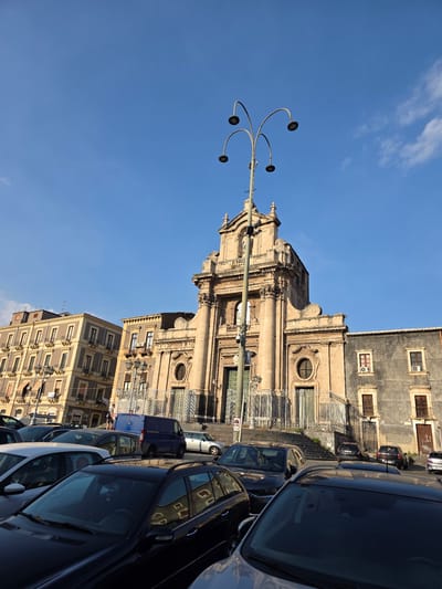 This is a low-angle, eye-level shot of the Piazza Duomo in Catania, Italy, on a clear day with a bright blue sky and a few wispy clouds. The focal point is the facade of the Catania Cathedral, a grand baroque structure made of pale stone, with a large central entrance, ornate columns, and decorative carvings. To the left of the cathedral, a multi-story yellow-brown building with numerous windows and balconies is visible. To the right of the cathedral, a dark gray building with a rough texture and fewer windows is present.
In the foreground, a parking lot is filled with various cars, including several dark-colored sedans and station wagons. A blue van is parked near the entrance of the cathedral. A tall, multi-headed street lamp with four lamps stands prominently in front of the cathedral. The steps leading up to the cathedral entrance are lined with decorative metal railings. There are no visible people in the immediate foreground, but a few figures can be seen on the steps of the cathedral in the mid-ground. The lighting suggests it is late afternoon or early evening, with shadows cast across the scene.