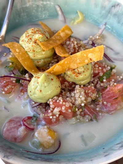 The image displays a close-up, overhead view of a prepared food dish presented in a light blue ceramic bowl with a textured, slightly uneven rim. The dish features a creamy, light green-tinted liquid base, likely a coconut milk or leche de tigre. Submerged and partially submerged ingredients include: diced pinkish-red segments, consistent with fish or seafood; numerous small, white, translucent grains resembling quinoa; thinly sliced red onions; and small pieces of diced green and yellow vegetables.
Three distinct dollops of a pale green, smooth purée, possibly an avocado or herb cream, are strategically placed on top of the solid ingredients, each lightly sprinkled with reddish-orange spice flakes. Several crisp, golden-yellow, elongated strips, resembling fried wonton or tortilla strips, are arranged across the dish, intersecting the green purée dollops. A metallic spoon handle is visible along the upper left edge of the bowl, partially submerged in the liquid.
The scene is a still life of a culinary presentation, likely captured in a restaurant or dining setting in Aldoar, Foz do Douro e Nevogilde, Portugal. No other people, actions, or specific environmental details beyond the dish and bowl are discernible.