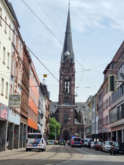 The image shows a street scene in Saarbrücken, Germany, with a view toward the Ludwigskirche church. The view is framed by buildings on either side of the street. Power lines stretch across the sky. Several police vehicles are parked along the right side of the street with flashing lights. Some pedestrians are near the vehicles. Buildings on the left display signs and architectural details. Graffiti is visible on concrete structures at the bottom of the frame. A bicycle lies on the ground.