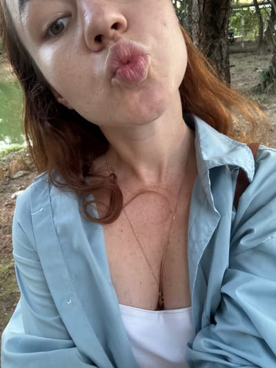 This is a close-up, vertically oriented selfie of a person with auburn hair, outdoors. The subject is leaning in, puckering their lips for a kiss. Their face is partially visible, with one eye looking towards the camera. The person is wearing a light blue, open collared shirt over a white top, and a delicate gold necklace. The background appears to be a natural setting with trees and possibly water, suggesting a park or garden. The lighting indicates it is daytime, possibly morning or afternoon, with soft, natural light. There is no visible text in the image. The overall mood is playful and intimate.