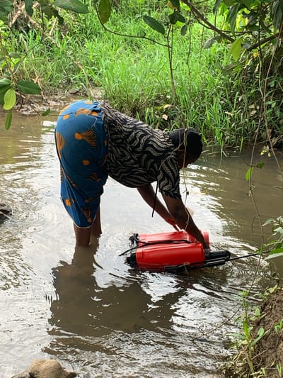 The image shows a person in a stream in Agban, Nigeria. The individual is crouched in the water wearing a dark patterned shirt and blue patterned shorts. They are interacting with a large, orange object that appears to be a container or piece of equipment partially submerged in the stream. The surrounding environment includes lush green vegetation on the banks of the stream, with trees and grass visible. The water is murky.