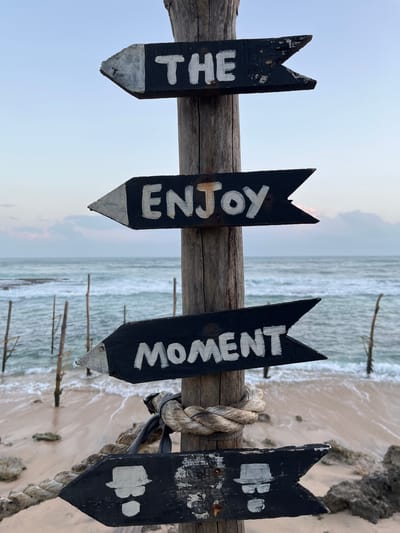 Four black wooden directional signs are attached to a rough wooden post, with white lettering on each. The top sign points left and reads "THE." The second sign from the top points left and reads "ENJOY." The third sign from the top points left and reads "MOMENT." The bottom sign points left and contains two stylized images of figures wearing hats and sunglasses. A thick rope is tied around the post below the "MOMENT" sign.
The signs are situated on a sandy beach with rocks and shallow water. In the background, the ocean is visible with waves and several thin, vertical wooden poles protruding from the water. The sky above is pale blue with some clouds on the horizon. This scene is located in Koggala, Sri Lanka, known for its stilt fishermen.