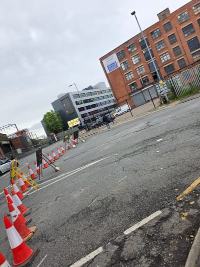 The image presents an urban street scene under an overcast sky, identified as likely being in the Ardwick area of Manchester, United Kingdom. This identification is based on a large white banner displayed on the prominent red-brick building, which reads "UNIVERSAL BUILDING" and "Belview," accompanied by the Manchester area code phone number "0161 236 0055" and website "www.belview.com."
In the foreground and extending into the mid-ground, an asphalt road runs diagonally from the lower left to the upper right. The road surface exhibits signs of wear, including cracks, patches, and minor debris. A series of red and white traffic cones, some upright and some leaning or toppled, are arranged along the left edge of the road, interspersed with yellow metal barriers. A broom with a long dark handle is propped against one of the cones in the mid-ground. White and yellow painted lines mark the road.
In the mid-ground, a solitary individual, dressed in a dark jacket, blue jeans, and white trainers, walks away from the camera on the asphalt, moving towards the right side of the frame.
On the right side of the street, the aforementioned five-story red-brick building with numerous dark-paned or boarded windows stands prominently. A tall, dark grey lamppost is positioned in front of this building. To its left, a chain-link fence separates the road from a paved area where a white van is parked.
Further to the left, behind another fence, a modern multi-story grey building with horizontal bands of multi-colored windows (yellow, green, blue, purple) is visible. A yellow billboard sign, partially visible, reads "GET MORE KNOWLEDGE YOUR WAY..." and is attached to this building. A classic red British telephone booth is situated near this modern structure, and a dark grey car is parked beside it.
In the far left background, railway infrastructure, including catenary wires and supporting structures, is present above a low-rise brick building, indicating proximity to train lines. A silver car is partially visible in the bottom-left corner of the frame, near the line of traffic cones. The lighting across the scene is flat and diffused, consistent with heavy cloud cover.