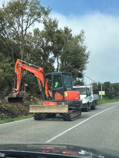 The image shows an orange Kubota excavator parked on the side of a road in Cisterna di Latina, Italy. Behind the excavator is a white truck with tires piled in its bed. Trees are present alongside the road. A utility pole with a sign is visible in the background. The photo appears to be taken from inside a vehicle.