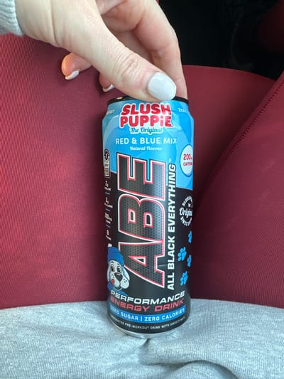 A light-skinned hand with white manicured fingernails holds the top portion of a can of ABE (All Black Everything) Performance Energy Drink. The can is labeled "SLUSH PUPPIE The Original Red & Blue Mix" and indicates "200mg CAFFEINE," "ZERO SUGAR," and "ZERO CALORIES." A cartoon dog mascot wearing a blue hat is depicted on the lower front. The can rests against a dark red, ribbed fabric surface, likely upholstery, with a lighter grey knitted fabric visible beneath it on the left. The background is indistinct, and the specific location within the city of Wealden, United Kingdom, cannot be determined from this image.