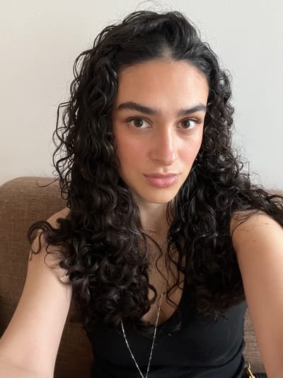 A young woman is posing for a selfie. She has dark curly hair, dark eyebrows, and light skin. She is wearing a black top. She appears to be sitting on a brown upholstered chair or sofa. The background is a plain, light-colored wall. The photo does not contain sufficient information to determine if the location is in the city of Lyon, France.