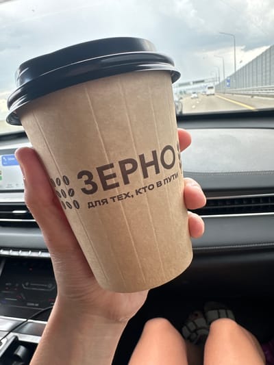 In Maryanskaya, Russia, a person is holding a disposable coffee cup with a black lid inside a vehicle. The cup has vertical ridges and text in Cyrillic script, which appears to be a brand name or slogan. The background shows a highway scene with other vehicles, streetlights, and a sound barrier. The interior of the car, including the dashboard and a portion of the passenger's legs and feet wearing sandals, is also visible. The lighting suggests it is daytime, with a cloudy sky.