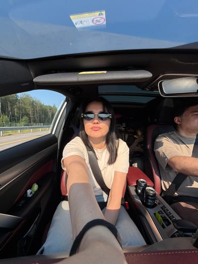 The image shows three individuals inside a car. A woman in the front passenger seat is looking at the camera, wearing sunglasses and a light-colored t-shirt. A man is driving in the driver's seat, partially visible. In the back seat, another person is seen wearing glasses. The car's interior features red and black upholstery, a sunroof, and a console with two cans of drinks. Outside the car, trees and a road are visible, indicating it is likely a scenic route. The location of Bolshiye Dvory, Russia is not discernible from the image alone.