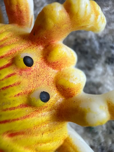 A close-up, macro photograph shows a yellow and orange plastic toy. The toy has two black, oval eyes embedded in its surface, which is textured and appears slightly fuzzy. Red stripes are painted across the yellow body of the toy. The background is out of focus and appears to be a rough, gray surface. There is no indication of the city of Ridabu, Norway, or any other specific location.