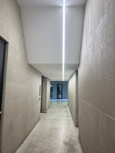 A long, clean, and modern corridor or passageway is depicted, characterized by a minimalist architectural style. The scene is static and devoid of people or discernible activity.
The floor, ceiling, and the right-hand wall of the corridor are uniformly covered in light grey, rectangular tiles with a subtle textured finish. These tiles create a consistent visual pattern throughout much of the space. In contrast, the left-hand wall is smooth, matte white, providing a clean break from the tiled surfaces.
A prominent feature is a bright, linear LED light strip embedded horizontally along the upper section of the white left wall, extending into the distance and illuminating the passageway. The light appears artificial, suggesting an interior space or a covered outdoor area.
The perspective looks down the length of the corridor, which recedes towards a darker background. On the right side of the passage, further back, there is a raised, elongated structure or platform, also clad in the same light grey tiles. At its base, a small, light-colored, crumpled object, possibly a sack or cloth, rests on the floor.
At the very end of the corridor, the space opens up to a darker area, revealing what appears to be glass partitions or windows that reflect light. Through this glass, faint white text is visible, including what looks like "do nion" and possibly a letter "A" or the number "4" on a dark surface. In the immediate foreground, at the bottom of the frame, there is a dark, rectangular opening, likely a doorway or access panel.
The overall impression is one of quiet order and functional design within a modern setting, consistent with contemporary architecture found in locations like Genève, Switzerland.