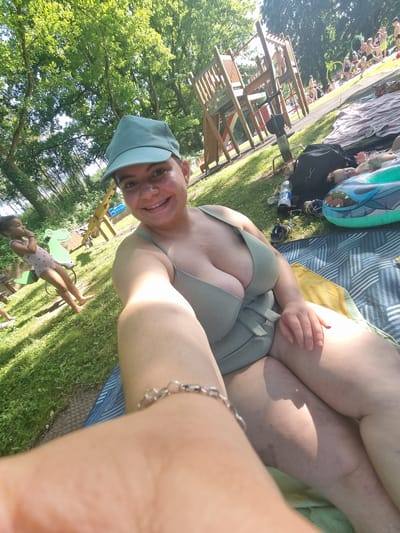 The image shows an individual wearing a light green bikini top and a matching cap, lying on a striped towel on grass. A silver bracelet is visible on the individual's wrist. In the background, there's a playground structure with wooden components, other people, and inflatables. The location appears to be a park or recreational area. The city of Saarbrücken, Germany, is not discernable from the image.